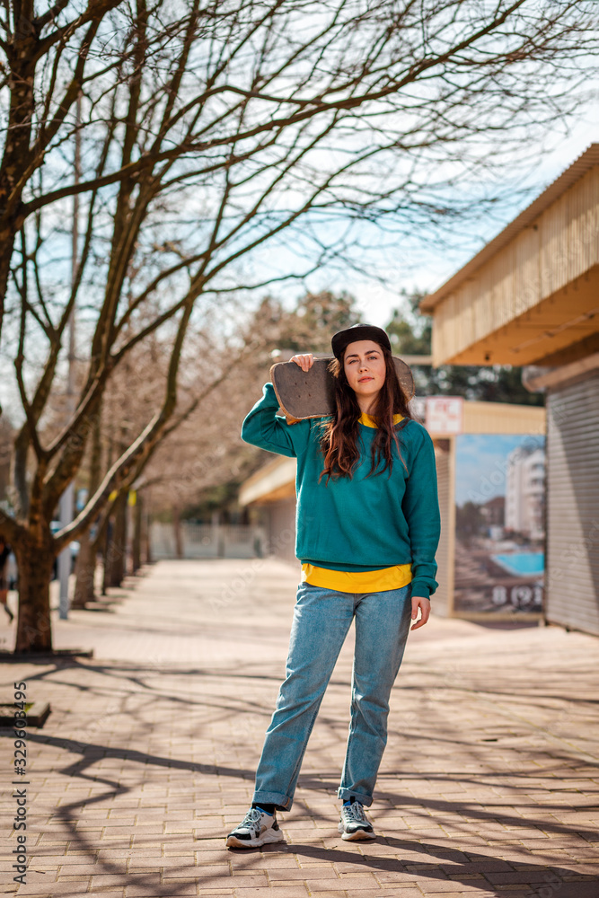 Fototapeta premium A young Caucasian hipster woman poses with a skateboard over her shoulders. In the background, an alley. Vertical. Concept of sports lifestyle and street culture