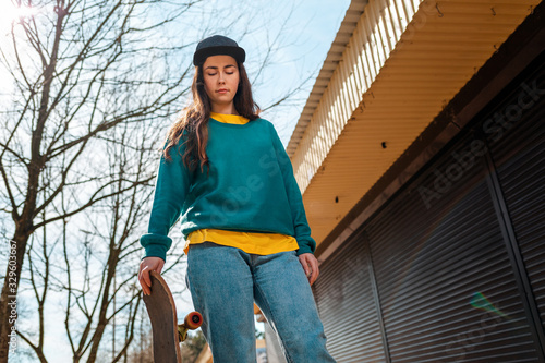 A portrait of young Caucasian hipster woman poses with a skateboard. In the background, an alley. Bottom view. Concept of sports lifestyle and street culture © _KUBE_