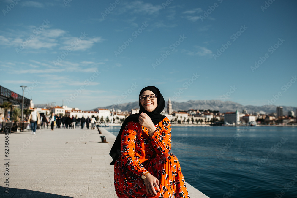 portrait of European Muslim women with hijab sitting on the stone beach with sea and port in the background. She is holding bouquet of yellow flowers.