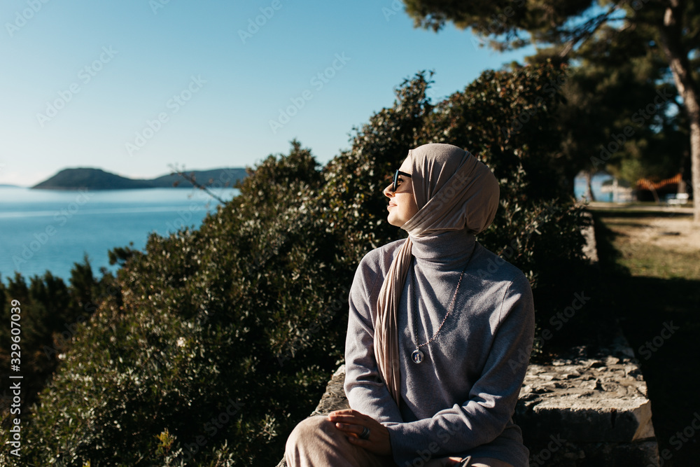 Fototapeta premium portrait of young European Muslim women with hijab sitting on the stone beach with sea and port in the background. She is happy and enjoying sun.