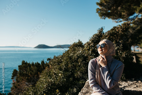 portrait of young European Muslim women with hijab sitting on the stone beach with sea and port in the background. She is happy and enjoying sun.