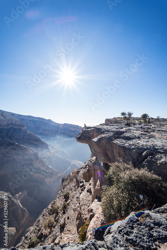 Man peeking over the cliff with the sun in the background at the Grand Canyon of Jebel Shams, Oman