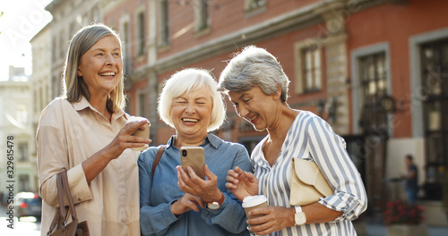 Three beautiful and cheerful Caucasian women with coffee to-go in hands laughing while scrolling photos on smartphone outdoor. Happy joyful grandmothers smiling while watching pictures on smartphone. photo