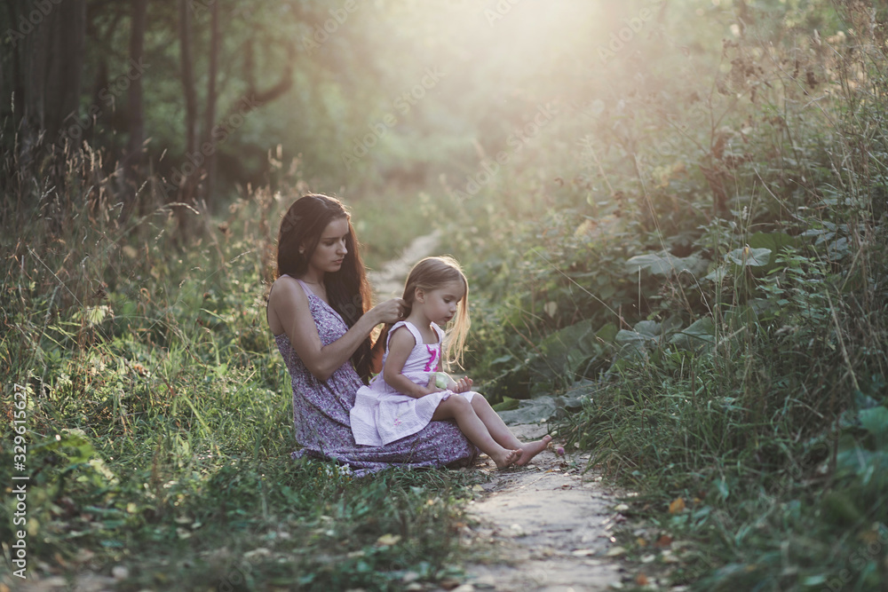 mother and child daughter in summer park on sunset. mom braids a little girl's hair mother and child daughter in summer park on sunset. mom braids a little girl's hair