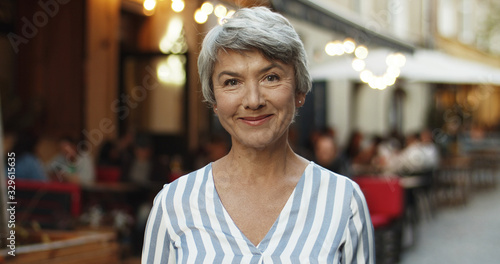 Portrait of beautiful senior Caucasian woman with grey hair looking straight at camera and smiling cheerfully outdoor in town. Close up of old lady with smile standing on street.