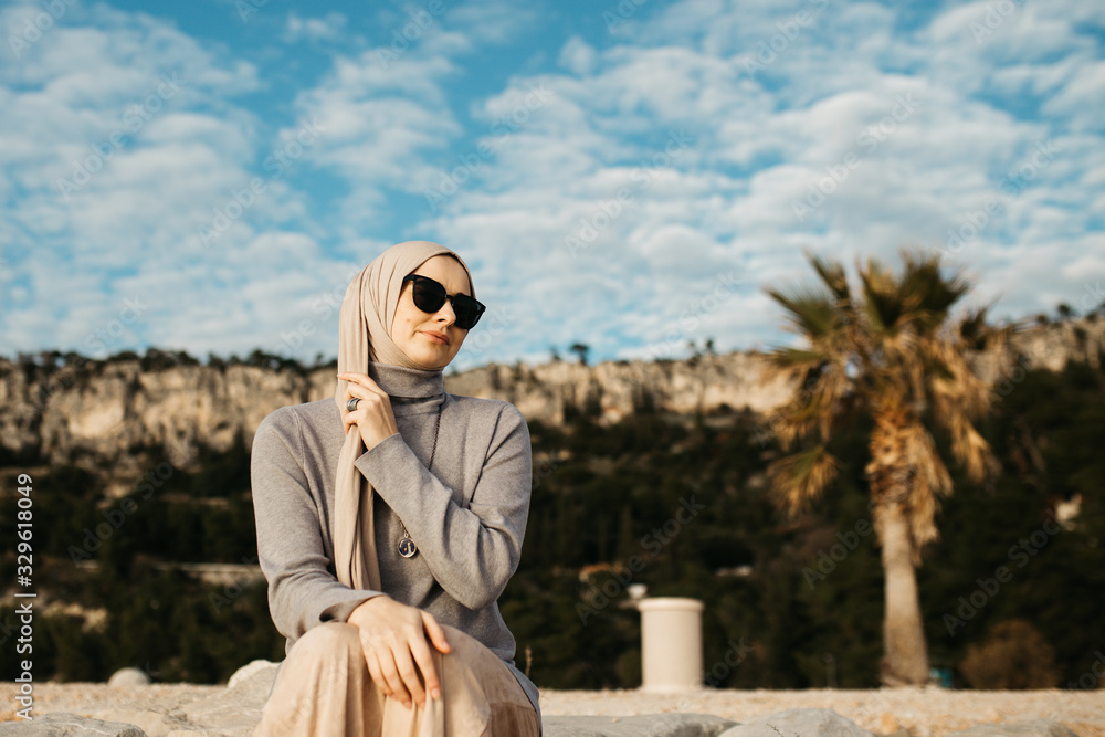 Fototapeta premium portrait of young European Muslim women with hijab sitting on the stone beach with sea in the background. She is happy and relaxed.