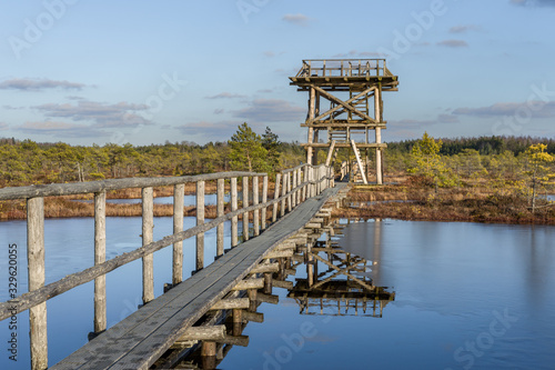 Wooden watch tower and pathway bridge over the lake in raised bog. Early spring, some pools are still frozen,  bonsai size pine trees. Bright day, blue sky and white clouds. Endla Nature reserve. photo