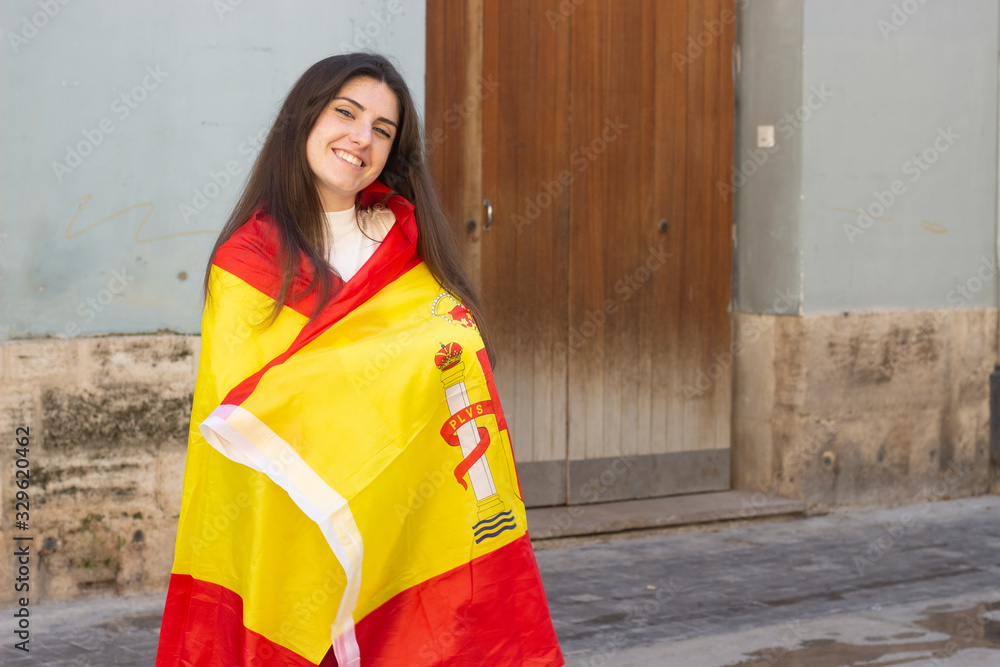 Fototapeta premium pretty caucasian young woman wrapped in a spanish flag, Spain, in the city, white top and red pants, dark air. Place for your text in copy space.