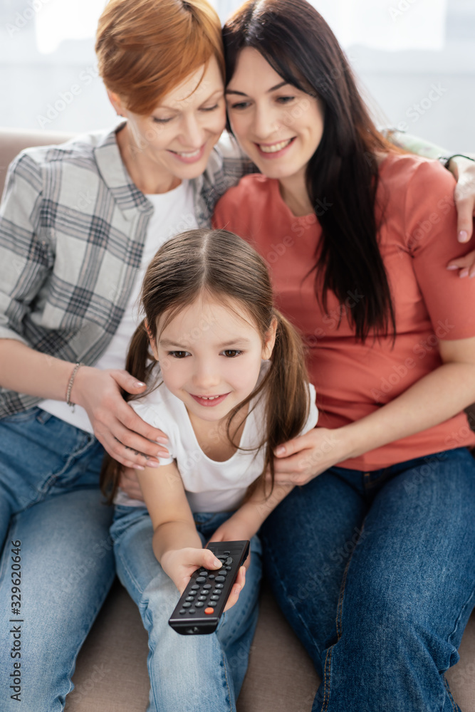 Selective focus of smiling child with remote controller watching movie near mothers on couch Selective focus of smiling child with remote controller watching movie near mothers on couch