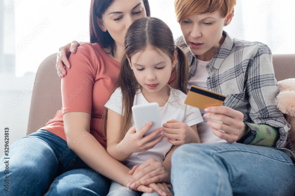 Same sex family hugging daughter while using smartphone and credit card on couch in living room