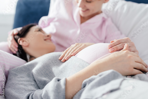 Selective focus of smiling woman looking at pregnant girlfriend in pajamas on bed