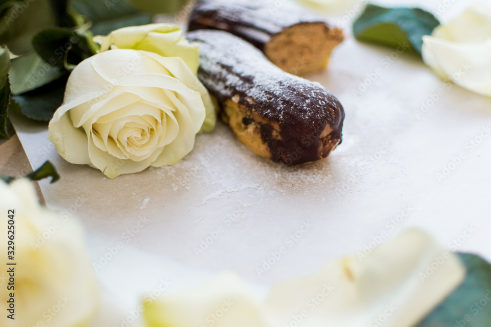 French eclairs with custard sprinkled with powdered sugar and white roses, petals, green leaves on light background