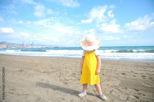 little girl in a yellow sundress on an empty beach on the background of the sea and the sky hid under a straw hat