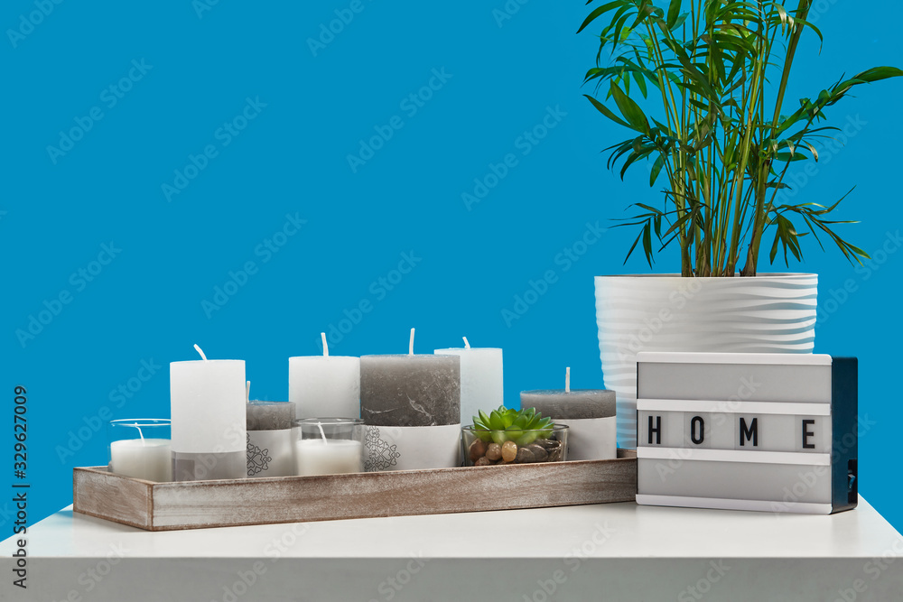 White table with green plant and succulent in pots, different sized candles in wooden stand, lamp with inscription home. Blue background. Close up White table with green plant and succulent in pots, different sized candles in wooden stand, lamp with inscription home. Blue background. Close up