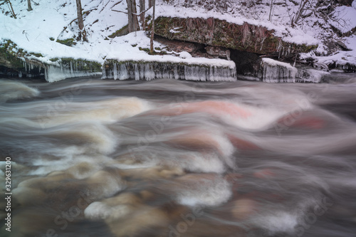 Long exposure photo of smooth rapid on the creek. River flowing in cold winter day. Rich of iron water is colored red. Estonia, North Europe.