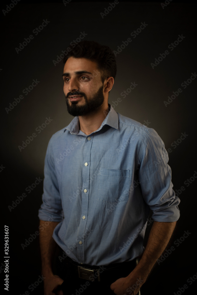 Fashion portrait of an young brunette Indian/European/Arabian/Kashmiri man in blue formal shirt and trousers in front of a black copy space background. Indian lifestyle and fashion photography.