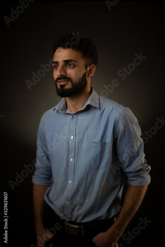 Fashion portrait of an young brunette Indian European Arabian Kashmiri man in blue formal shirt and trousers in front of a black copy space background. Indian lifestyle and fashion photography.