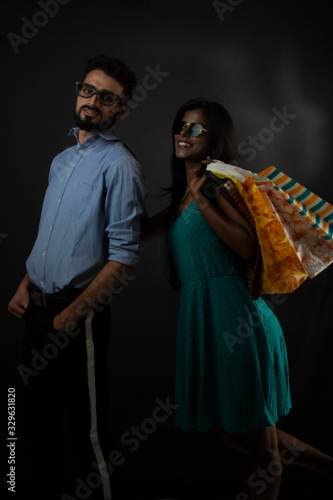 A dark skinned Indian/African girl and a Kashmiri/European/Arabian man in formal wear and glasses with shopping bag in front of black copy space studio background. Indian lifestyle.
