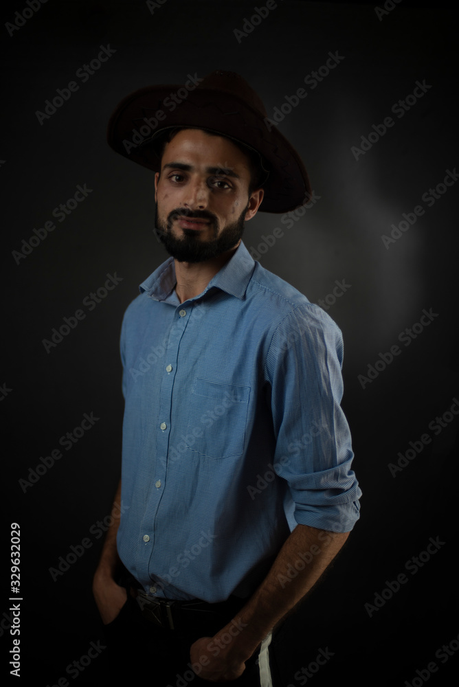 Portrait of young brunette Indian/European/Arabian/Kashmiri man in blue formal shirt and trousers with cowboy hat and pipe in front of black copy space background. Lifestyle and fashion photography.