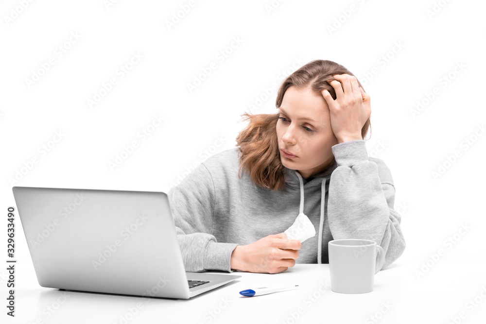 Young woman in hoodie sitting at table and holding napkin while reading about symptoms on internet using laptop Young woman in hoodie sitting at table and holding napkin while reading about symptoms on internet using laptop