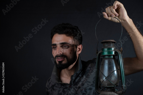 Portrait of young brunette Indian/European/Arabian/Kashmiri man in casual tee shirt holding a vintage lantern in black copy space studio background. Indian lifestyle and fashion.