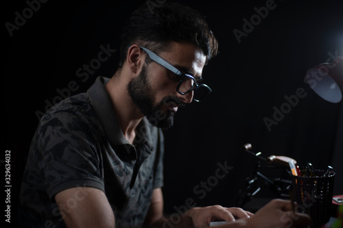 Portrait of young brunette Indian/European/Arabian/Kashmiri man in casual tee shirt with glasses studying in front of a table lamp in black copy space studio background. Indian lifestyle.