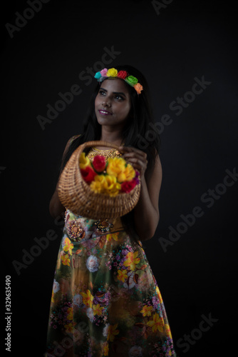 Fashion portrait of young dark skinned Indian Bengali brunette girl in floral dress with floral head band taking flowers in front of black copy space studio background. Indian fashion photography.
