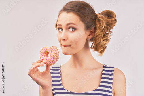A young attractive woman with a stylish hairstyle holds a scented donut in her hand and opens her eyes wide with a surprised expression. Close-up portrait on white background.
