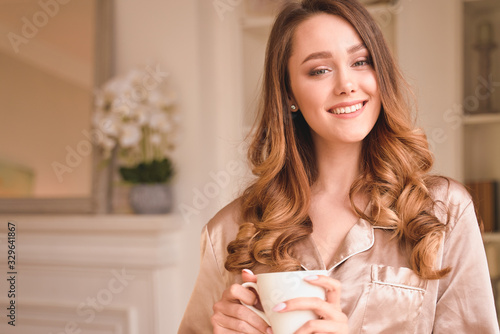 Sensitive young woman delights morning. Meeting sunrise, enjoyment and calmness concept, positive beginning of day. Lady has long blonde wavy hair, beautiful smile. Wearing nice fashionable pajama.