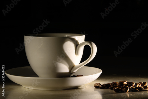 A white ceramic cup of coffee stands on a saucer. On a saucer and table are coffee grains. On the surface of the cup is a heart-shaped shadow. Black background. Concept - coffee lover, coffee break.