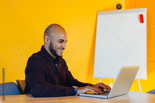 Smiling bearded african man using laptop at home, sitting on a wooden table. Concept of young people work mobile devices. Blurred window background, wide.