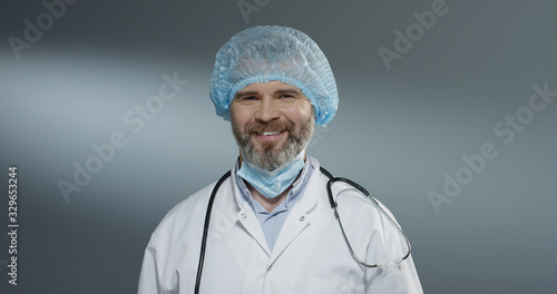 Portrait shot of the Caucasian handsome man doctor in blue hat taking off mask from his mouth and smiling cheerfully to the camera.