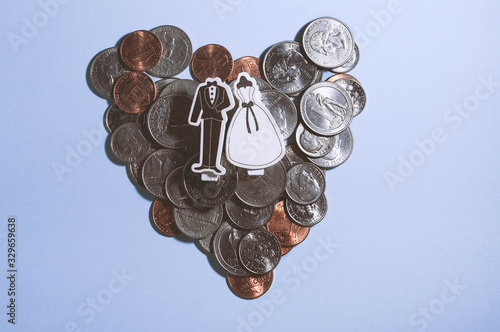 top view of decorative stylish bride dress and black groom tuxedo clamps together on pile heart-shaped of american dollars coins isolated on purple, expensive wedding concept