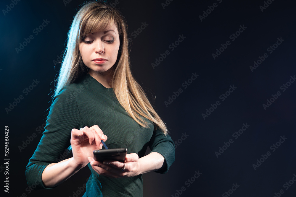 Woman on a black background with a phone in her hands. Girl holds a phone near the camera. Young woman works with a smartphone. Phone with a stylus. Concept - business woman. The student. PDA Woman on a black background with a phone in her hands. Girl holds a phone near the camera. Young woman works with a smartphone. Phone with a stylus. Concept - business woman. The student. PDA