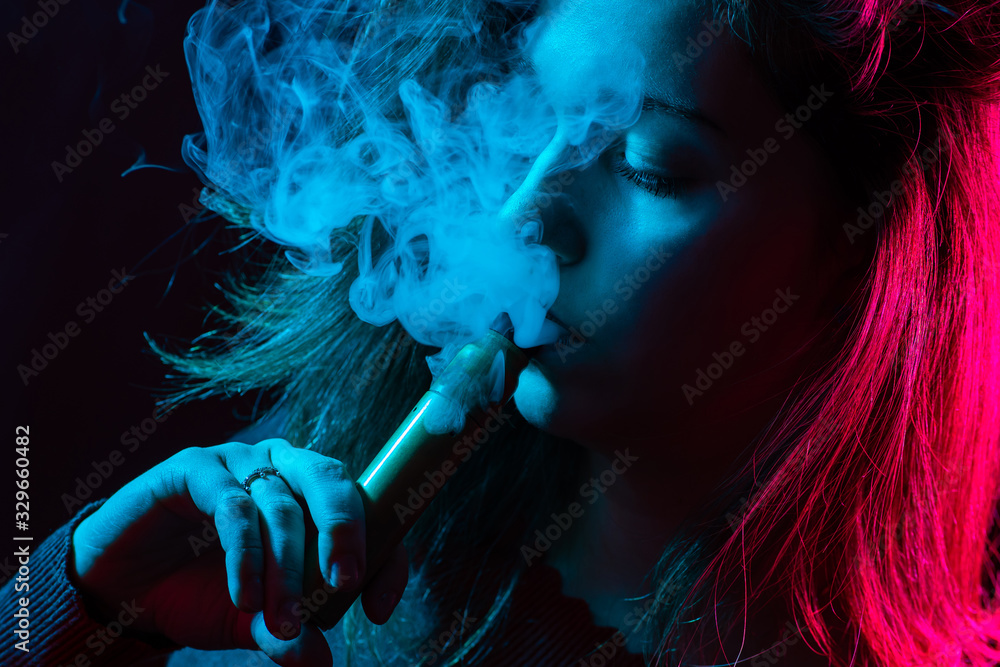 Girl releases smoke from her lips. Female student with an electronic cigarette in her hands. Face of a viper girl close-up. Woman brings a vape device to her mouth. Student vapit on a dark background Girl releases smoke from her lips. Female student with an electronic cigarette in her hands. Face of a viper girl close-up. Woman brings a vape device to her mouth. Student vapit on a dark background