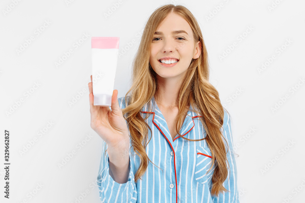 Obraz premium Portrait of happy beautiful young woman in pajamas standing isolated on white background applying face lotion