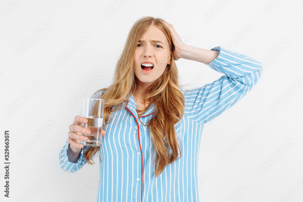 Portrait of a suffering young woman in pajamas holding a pill and a glass of water standing on a white background Portrait of a suffering young woman in pajamas holding a pill and a glass of water standing on a white background