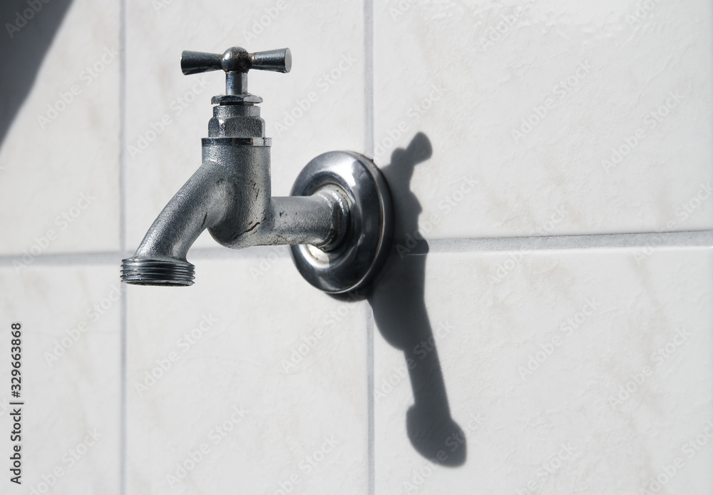 closeup of of silver hose bibb / stopcock on wall with white tiles, symbol for water wasting and dripping closeup of of silver hose bibb / stopcock on wall with white tiles, symbol for water wasting and dripping