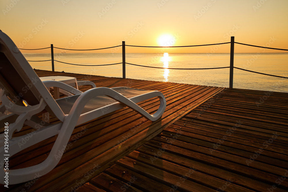 Naklejka premium Beach lounger stands on a wooden pier against the background of a calm sea and the rising sun