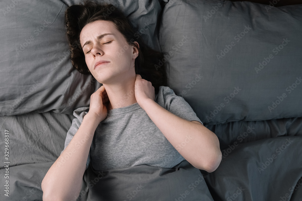 Top close up view upset young woman lying in bed in morning feels pain in neck after night sleep, awaken having painful sudden ache or stiffness, incorrect posture during sleep. Fibromyalgia concept Top close up view upset young woman lying in bed in morning feels pain in neck after night sleep, awaken having painful sudden ache or stiffness, incorrect posture during sleep. Fibromyalgia concept