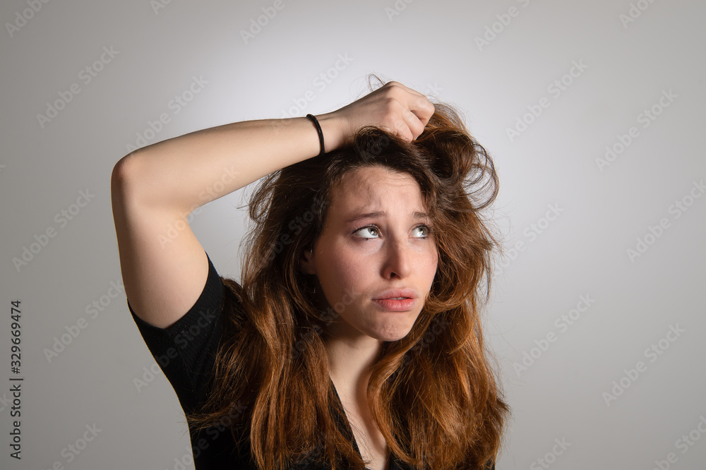 Fototapeta premium Portrait of frustrated young girl holding her long brown hair. Disappointed expression in female face. Isolated on grey background. Emotions...