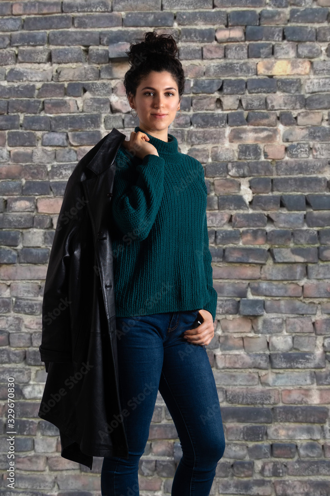 Naklejka premium Portrait of young attractive woman in sweater with leather jacket on her shoulder. Brunette female model with topknot hairstyle looking at camera. Old brick wall background...