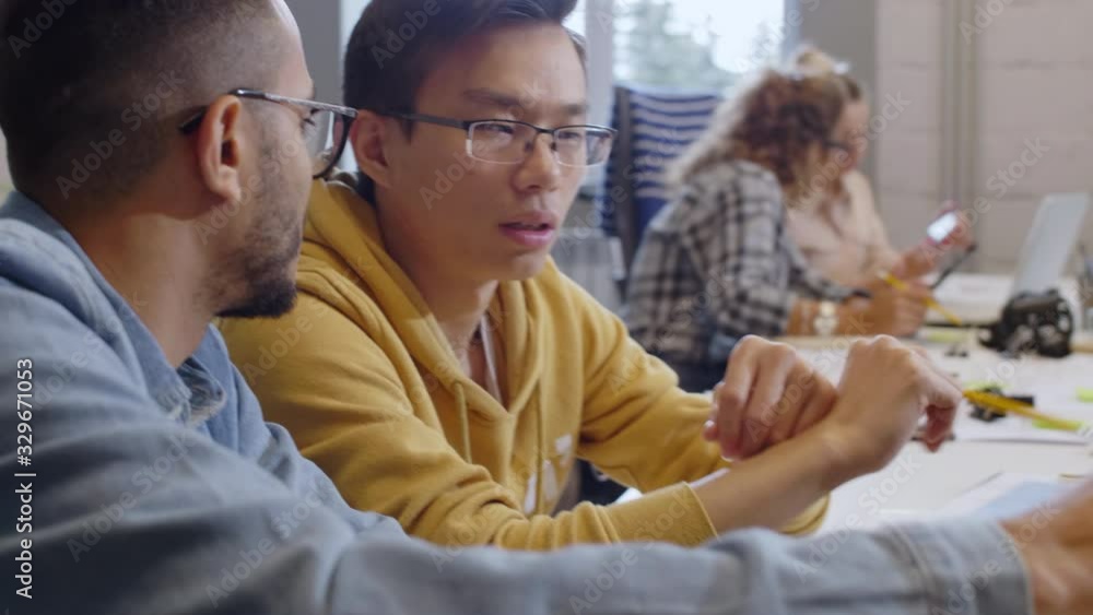 Tilt up shot of young Asian and middle eastern male colleagues pointing at laptop screen and discussing project at office desk Tilt up shot of young Asian and middle eastern male colleagues pointing at laptop screen and discussing project at office desk