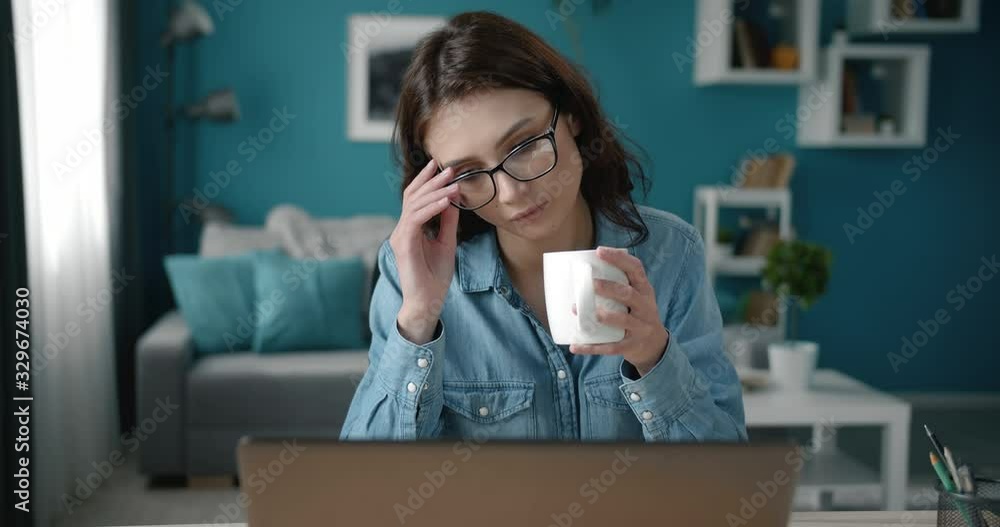 Attractive brown-haired woman in eyeglasses and jeans shirt drinking coffee and looking at computer screan at home. Serious lady with curly hair sitting at table and using laptop. Attractive brown-haired woman in eyeglasses and jeans shirt drinking coffee and looking at computer screan at home. Serious lady with curly hair sitting at table and using laptop.