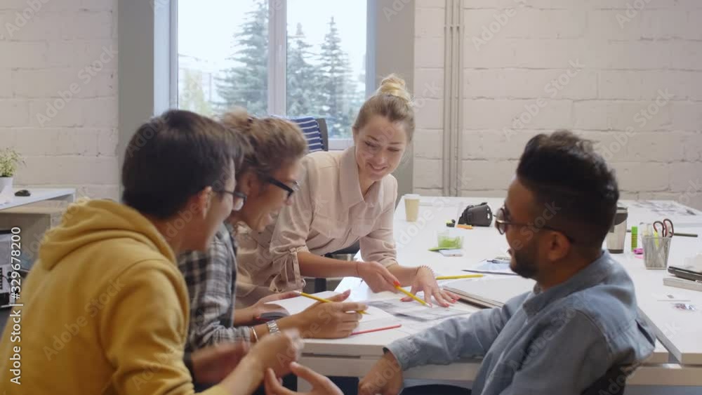 Group of young multiethnic male and female designers discussing sketches at office table, joking and laughing while working together on creative project Group of young multiethnic male and female designers discussing sketches at office table, joking and laughing while working together on creative project