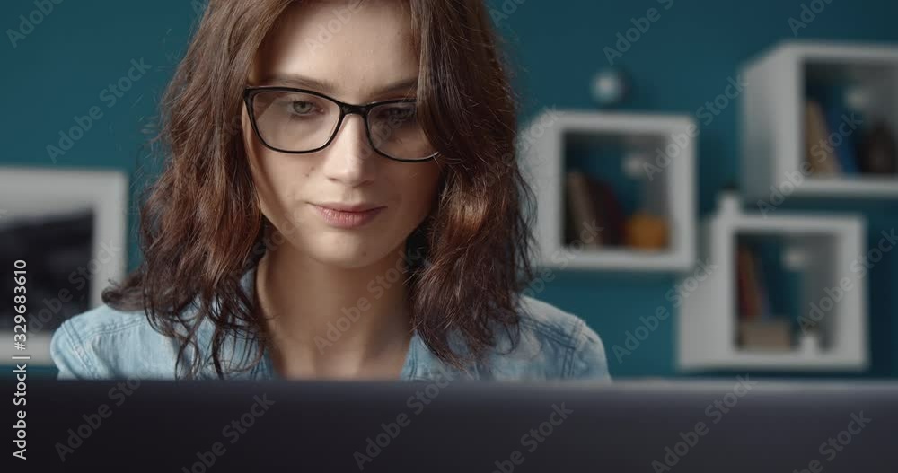Close up of attractive young girl in eyeglasses and jeans shirt typing on laptop while while sitting at desk at living room. Concept of freelance career. Close up of attractive young girl in eyeglasses and jeans shirt typing on laptop while while sitting at desk at living room. Concept of freelance career.