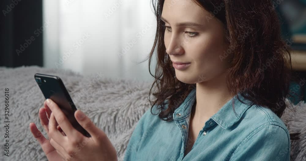 Attractive brunette in jeans shirt sitting on grey couch and smiling while looking at screen of mobile phone. Beautiful girl with brown hair reading message on smartphone at home. Attractive brunette in jeans shirt sitting on grey couch and smiling while looking at screen of mobile phone. Beautiful girl with brown hair reading message on smartphone at home.