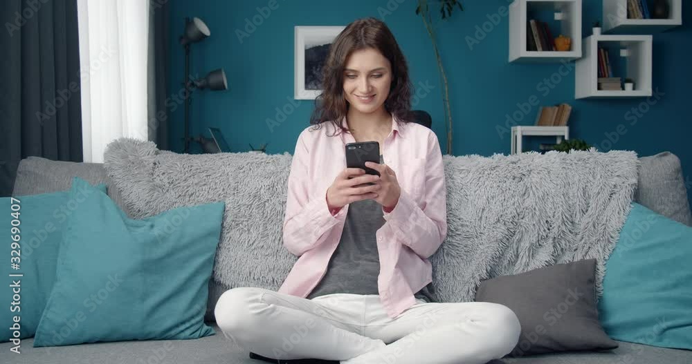 Beautiful young girl with curly hair on casual clothing sitting comfortably with crossed legs on sofa and using smartphone. Cheerful lady spending time with gadget at home. Beautiful young girl with curly hair on casual clothing sitting comfortably with crossed legs on sofa and using smartphone. Cheerful lady spending time with gadget at home.