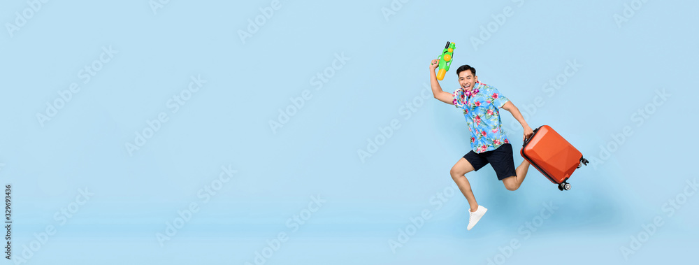 Jumping handsome Asian tourist man traveling with water gun and baggage during Songkran festival on banner blue background with copy space
