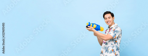Smiling handsome Asian man playing with water gun isolated on banner blue background with copy space for Songkran festival in Thailand and southeast Asia